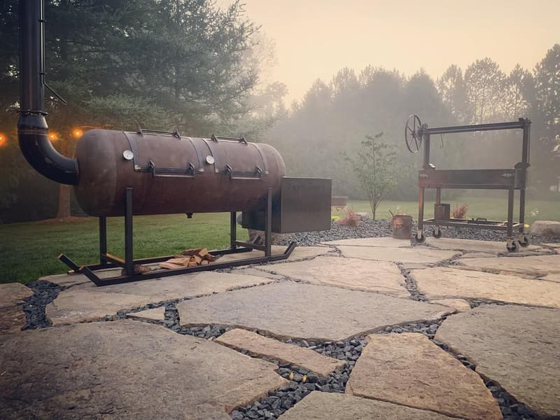 Offset smoker on a flagstone patio in morning fog, a live-fire grill behind it