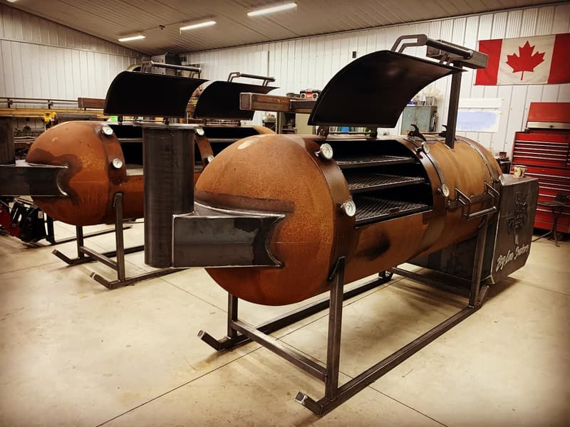 A row of rust-patina offset smokers with lids open inside the shop, Canadian flag on the wall
