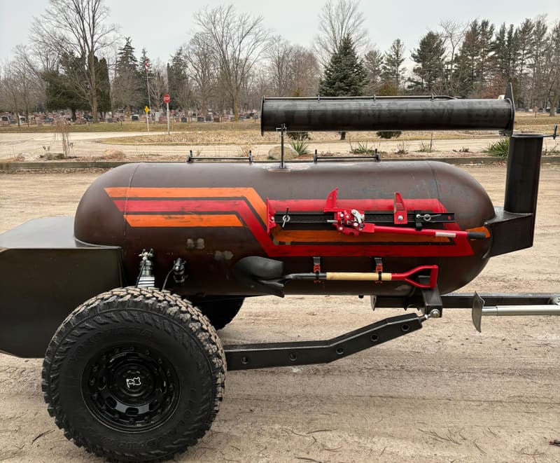 Trailer-mounted offset smoker with orange and red hot-rod stripes, off-road tires and a Hi-Lift jack